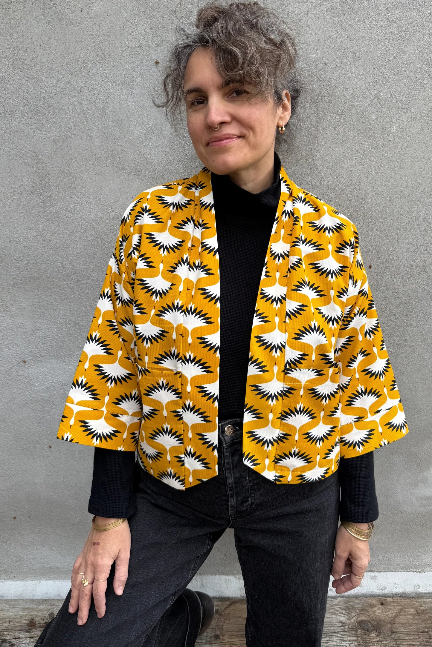 A woman wearing the Magnolia Jacket by Kazak in yellow corduroy Cranes print fabric, a short kimono style open jacket with 3/4 sleeves and large patch pockets. She is wearing it with a black top and black jeans and standing in front of a grey wall. 
