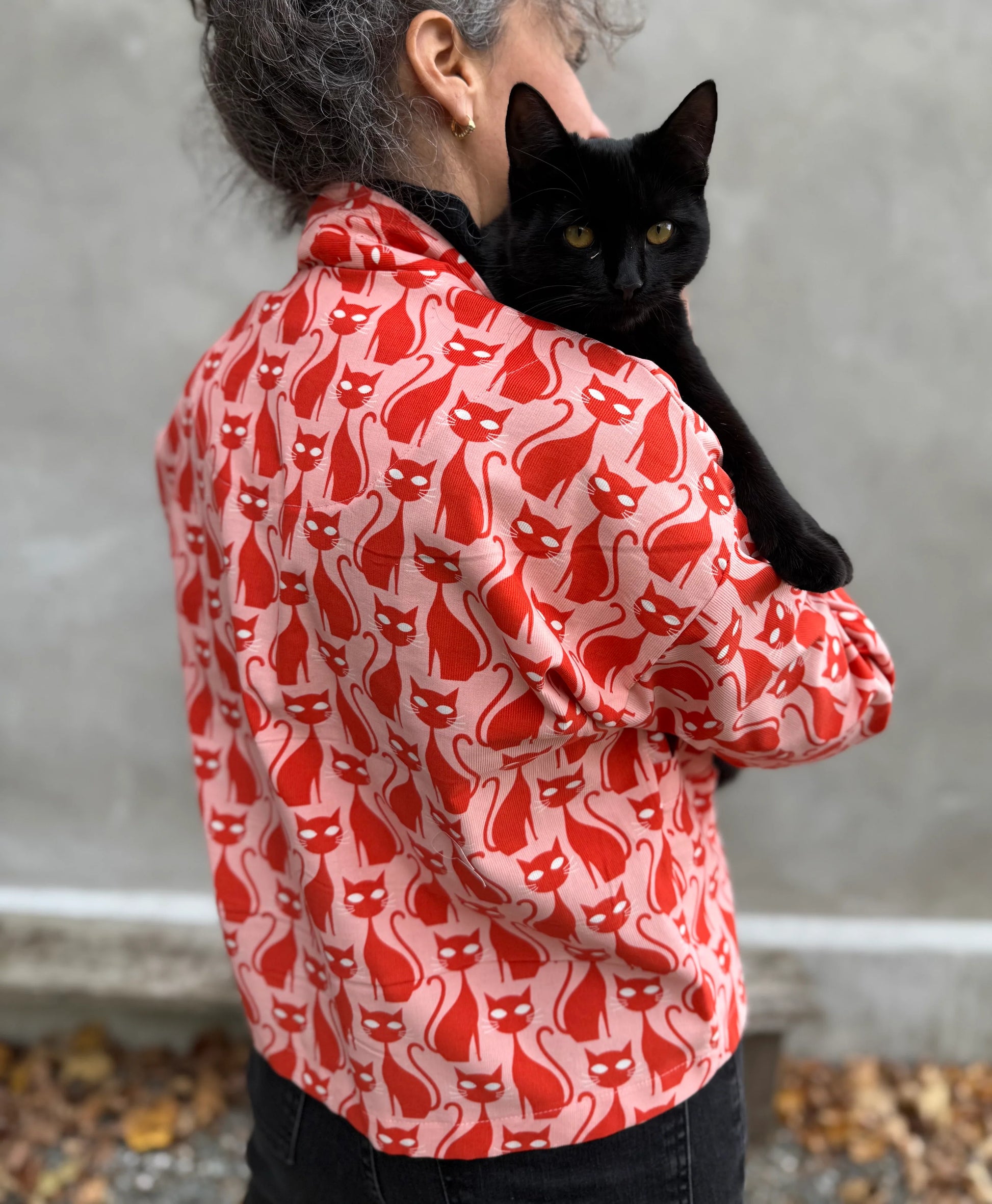 Back view of a woman wearing the Magnolia Jacket by Kazak in red corduroy Cats print fabric, a short kimono style open jacket with 3/4 sleeves and large patch pockets. She is wearing it with a black top and black jeans and holding a black cat while standing in front of a grey wall. 