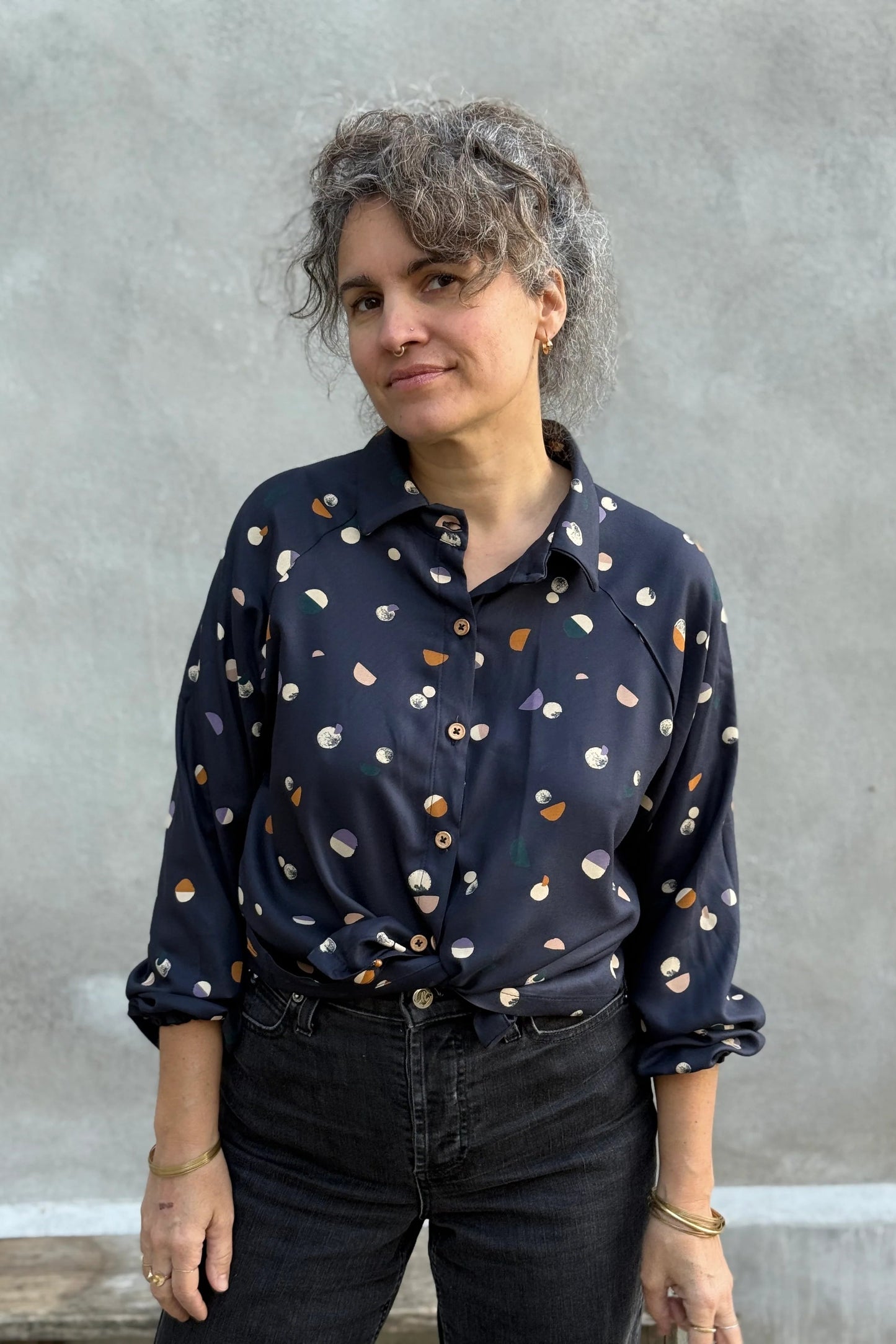 A woman wearing the Acer Blouse by Kazak in Midnight Moons, a raglan sleeved blouse with a classic collar, button front, and elastic cuffs. She is wearing it with black jeans and standing in front of a grey background. 