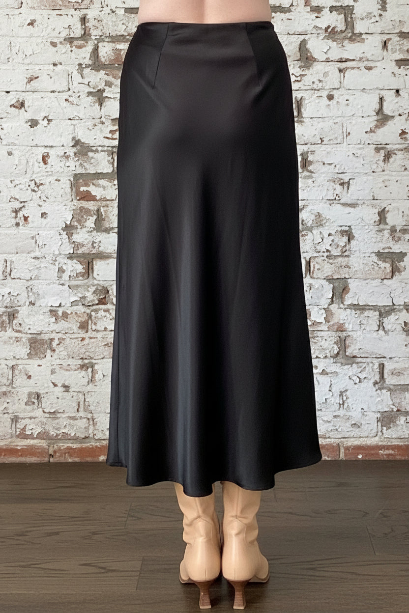Back waist down view of a woman wearing the Satin Skirt by Dotty in Black, a long bias cut A-line skirt with an elastic waistband. She is standing indoors in front of a white brick wall. 