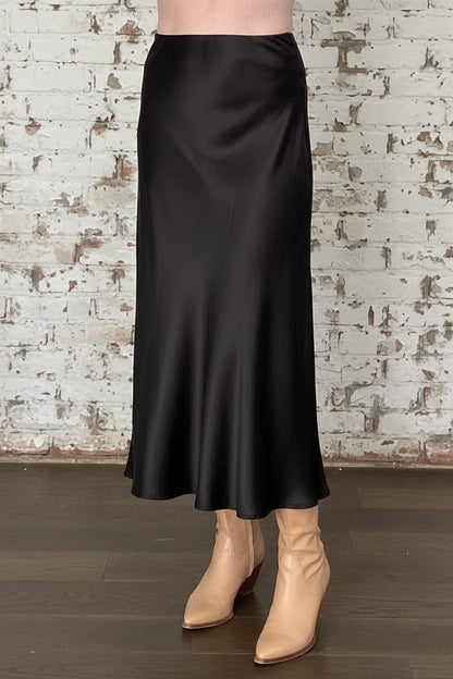 Waist down view of a woman wearing the Satin Skirt by Dotty in Black, a long bias cut A-line skirt with an elastic waistband. She is standing indoors in front of a white brick wall. 