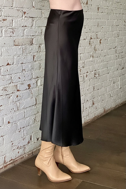 Side waist down view of a woman wearing the Satin Skirt by Dotty in Black, a long bias cut A-line skirt with an elastic waistband. She is standing indoors in front of a white brick wall. 