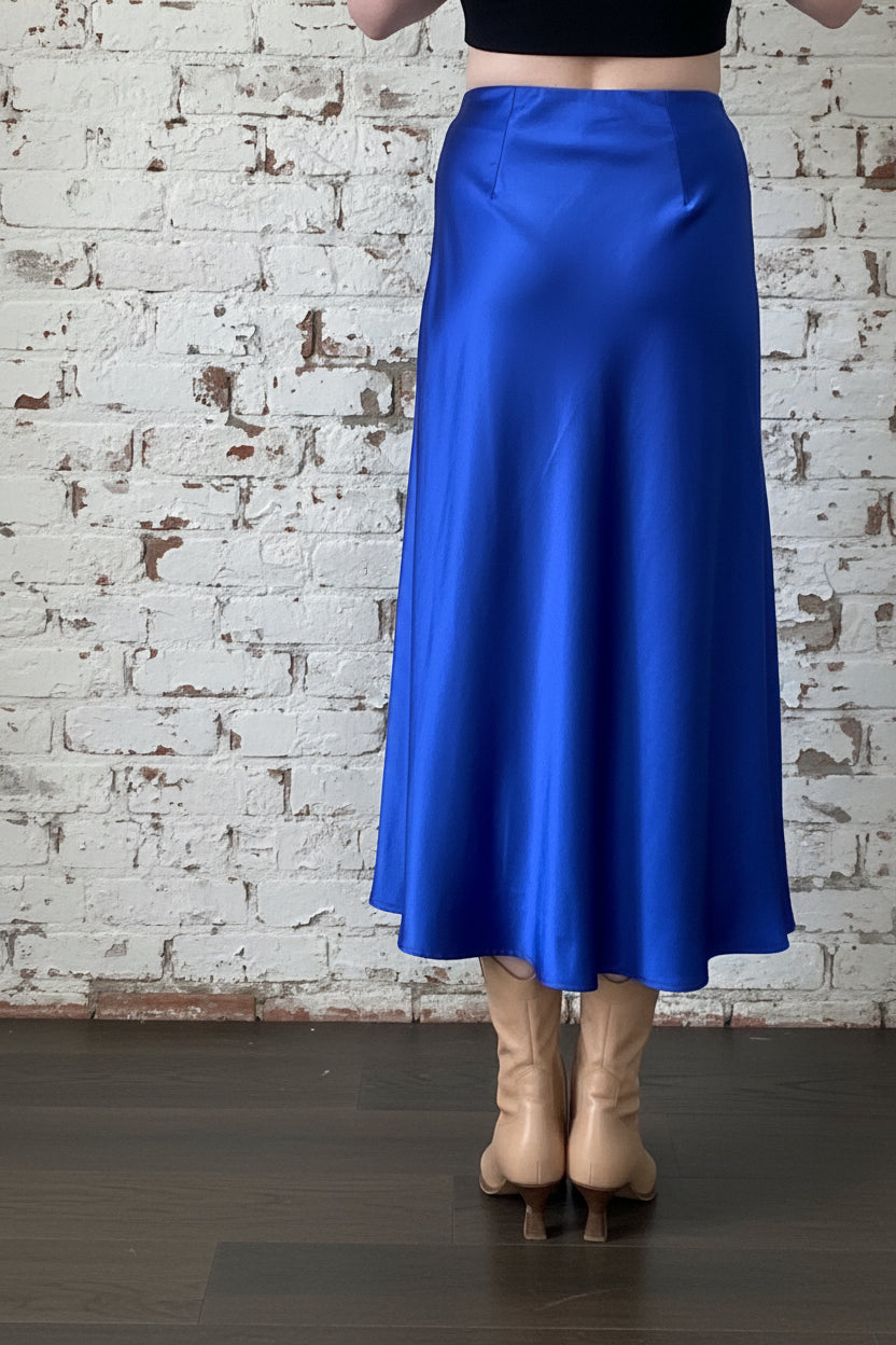 Back waist down view of a woman wearing the Satin Skirt by Dotty in Royal Blue, a long bias cut A-line skirt with an elastic waistband. She is standing indoors in front of a white brick wall. 