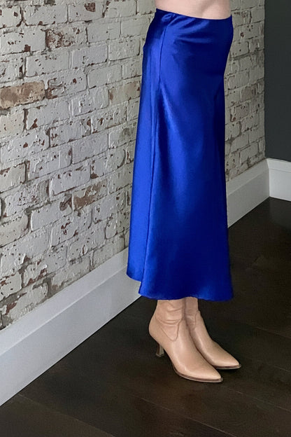 Side waist down view of a woman wearing the Satin Skirt by Dotty in Royal Blue, a long bias cut A-line skirt with an elastic waistband. She is standing indoors in front of a white brick wall. 