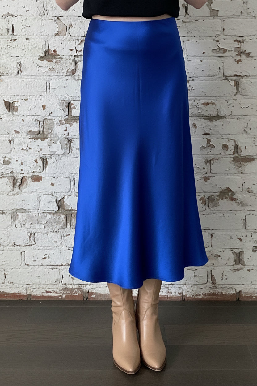 Waist down view of a woman wearing the Satin Skirt by Dotty in Royal Blue, a long bias cut A-line skirt with an elastic waistband. She is standing indoors in front of a white brick wall. 