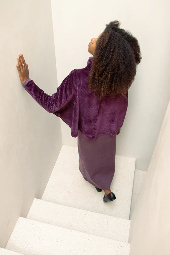 Back view of a woman wearing the Amsterdam Sweater by MAS in Amethyst, an oversized one-size top with a chimney collar, extended sleeves, and a cropped length, with a purple dress, standing in a white stairwell.