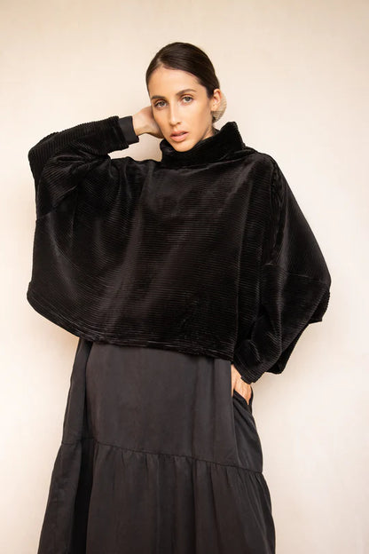 A woman wearing the Amsterdam Sweater by MAS in Black, an oversized one-size top with a chimney collar, extended sleeves, and a cropped length, with a black skirt, standing in front of a beige wall. 