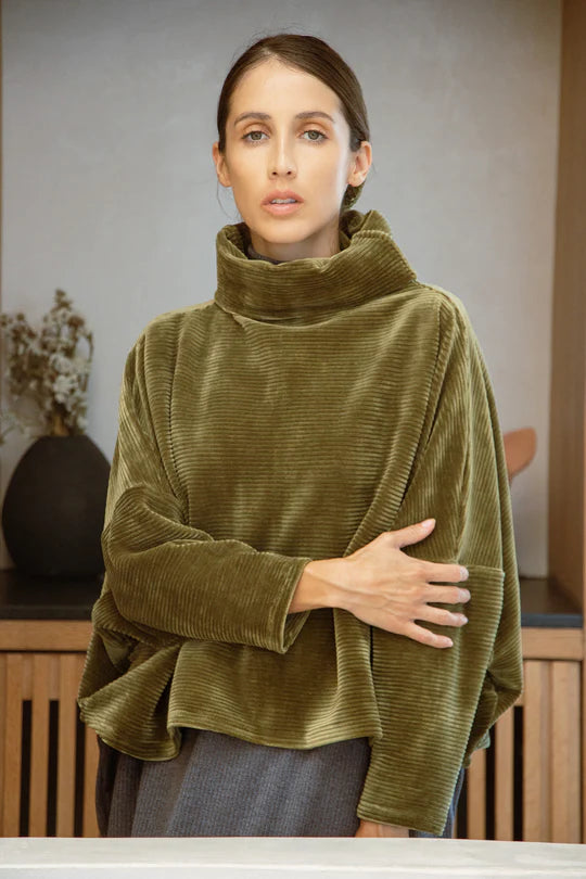 A woman wearing the Amsterdam Sweater by MAS in Moss, an oversized one-size top with a chimney collar, extended sleeves, and a cropped length, with blue trousers, standing in front of a wooden table. 