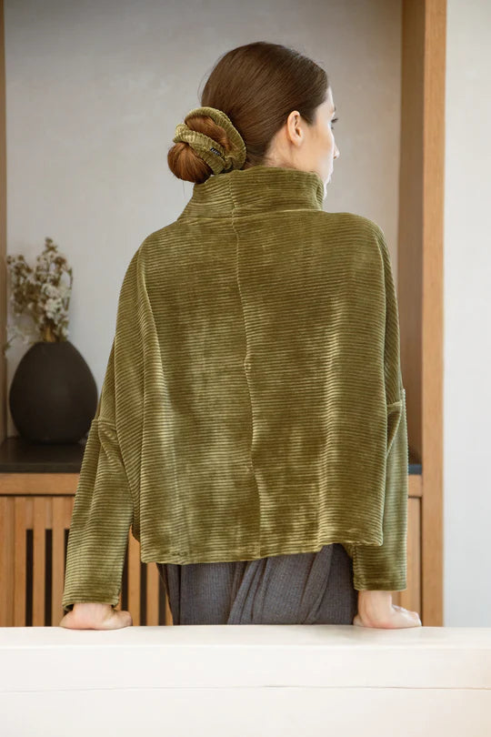 Back view of a woman wearing the Amsterdam Sweater by MAS in Moss, an oversized one-size top with a chimney collar, extended sleeves, and a cropped length, with a grey skirt, standing in front of a wooden table. 