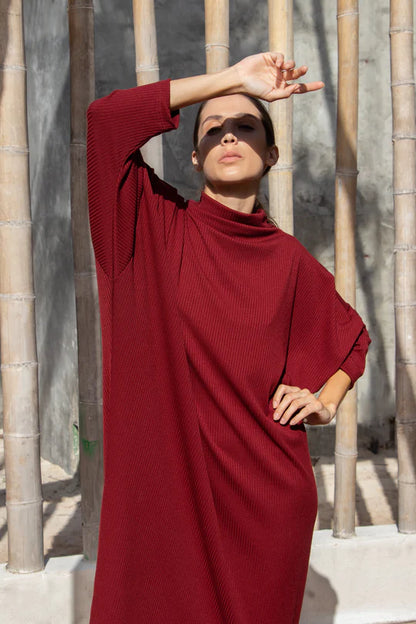 3/4 shot of a woman wearing the Frankfurt Dress by MAS in Ruby, standing outside front of a bamboo fence 