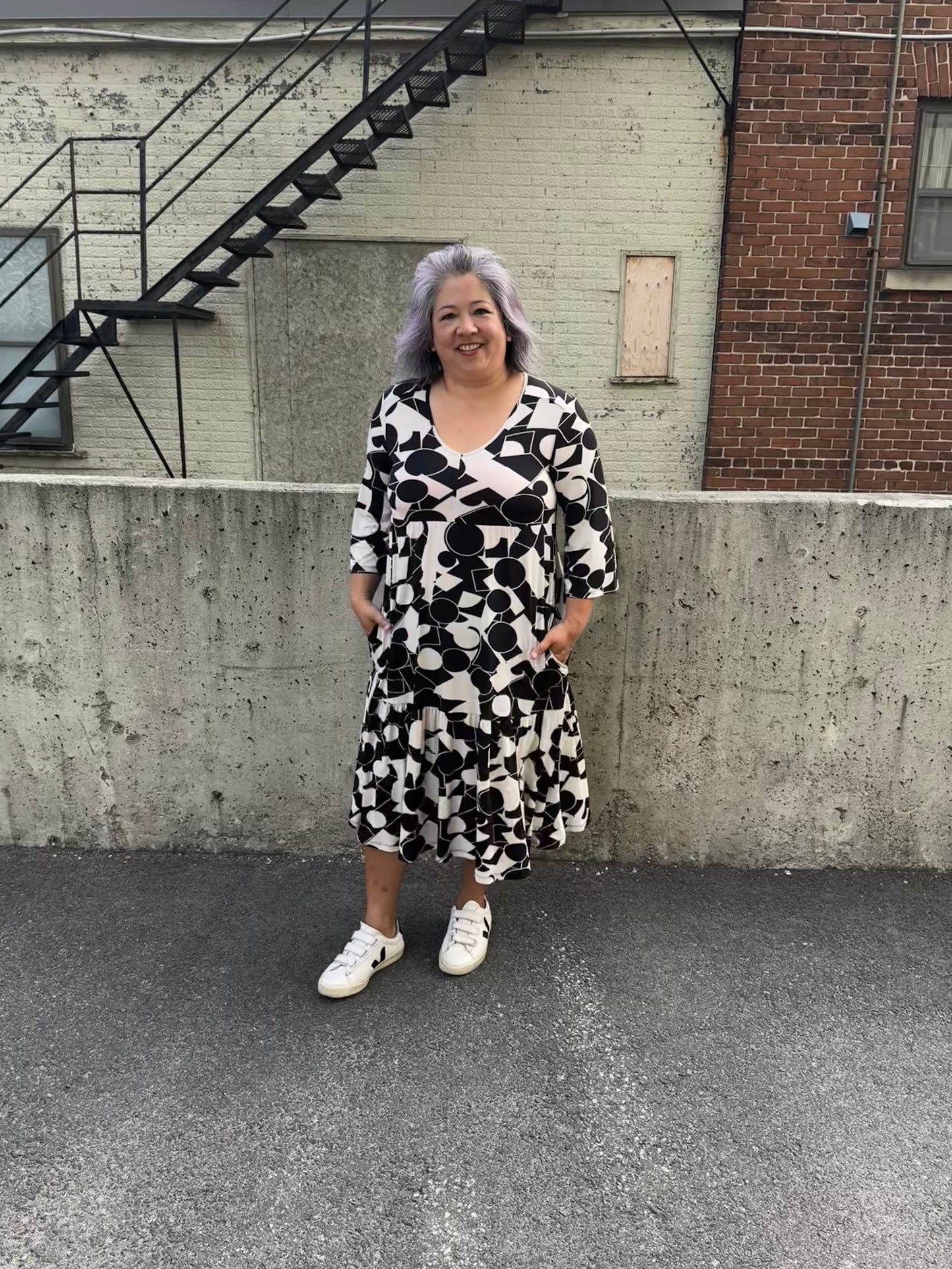 A woman wearing the Farah Dress by Pure Essence in Black/Ivory, a geometric print dress with a v-neck, 3/4-sleeves, baby doll shape, tiered midi length skirt, and front patch pockets. She is standing in front of a brick building. 