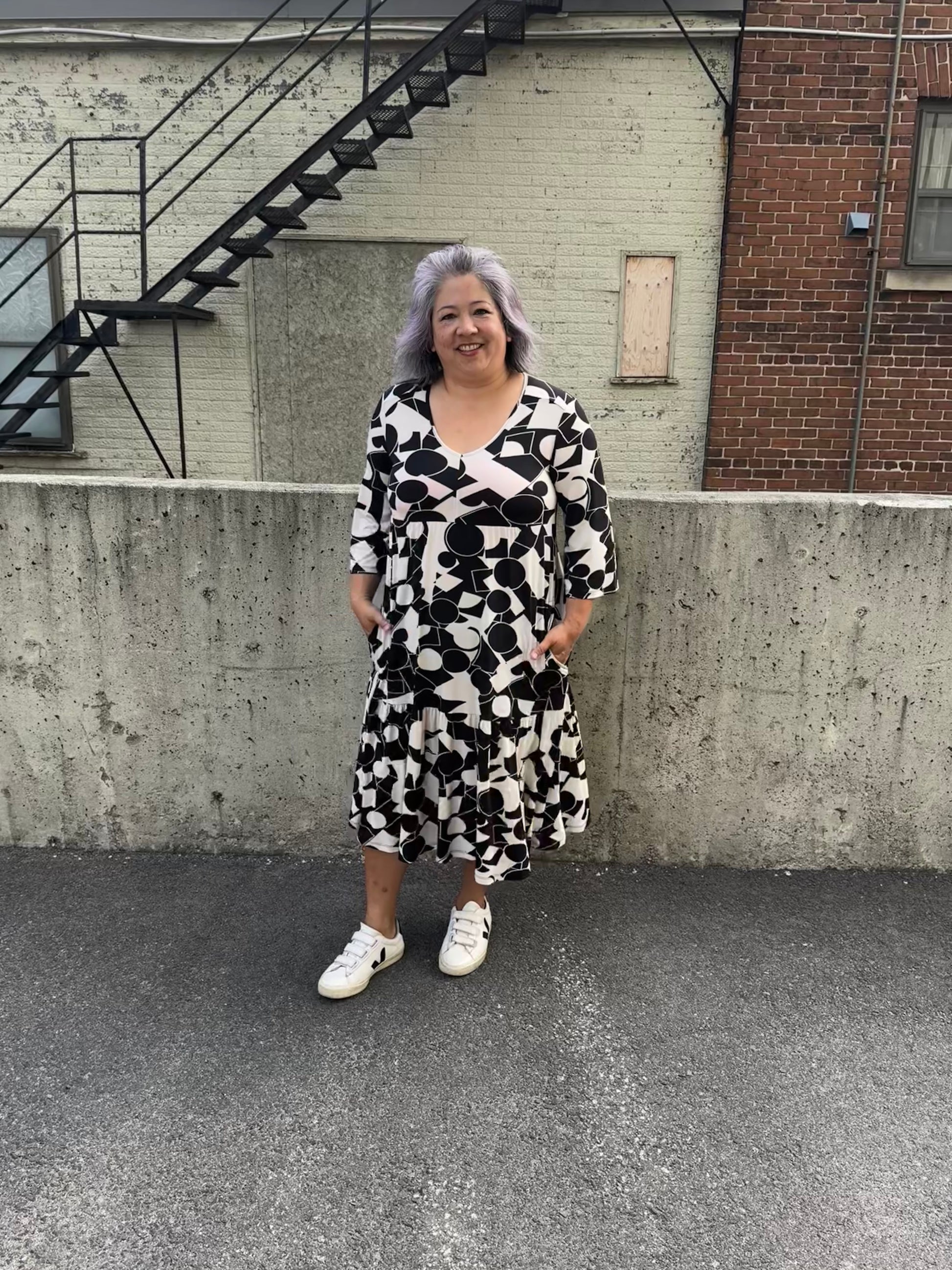 A woman wearing the Farah Dress by Pure Essence in Black/Ivory, a geometric print dress with a v-neck, 3/4-sleeves, baby doll shape, tiered midi length skirt, and front patch pockets. She is standing in front of a brick building. 