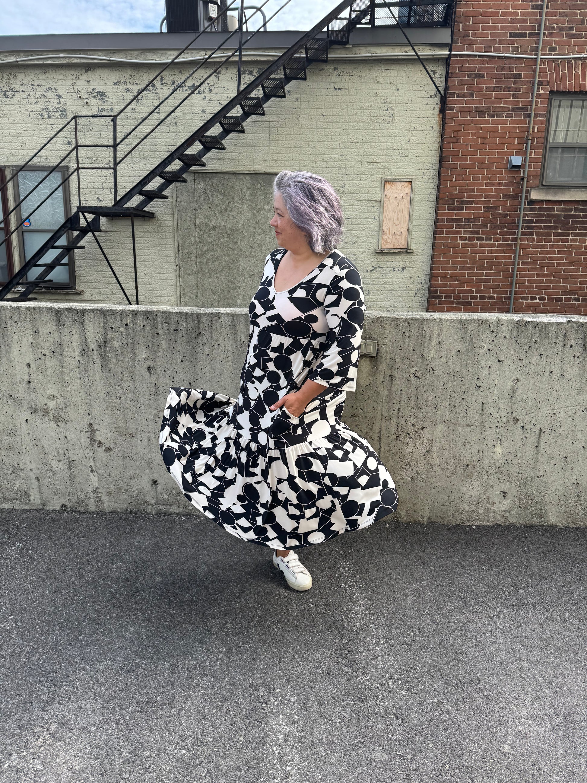 A woman wearing the Farah Dress by Pure Essence in Black/Ivory, a geometric print dress with a v-neck, 3/4-sleeves, baby doll shape, tiered midi length skirt, and front patch pockets. She is twirling in front of a brick building. 