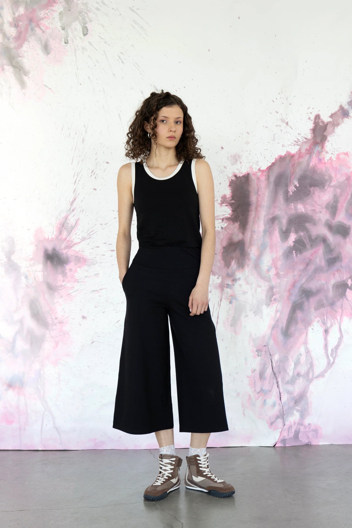 A woman wearing the Pissenlit Camisole by Eve Lavoie in Black/White, a black tank with white contrast piping at the neck and shoulders. She is wearing it with the Trille Trousers in Black and standing against a abstract pink and grey wall.