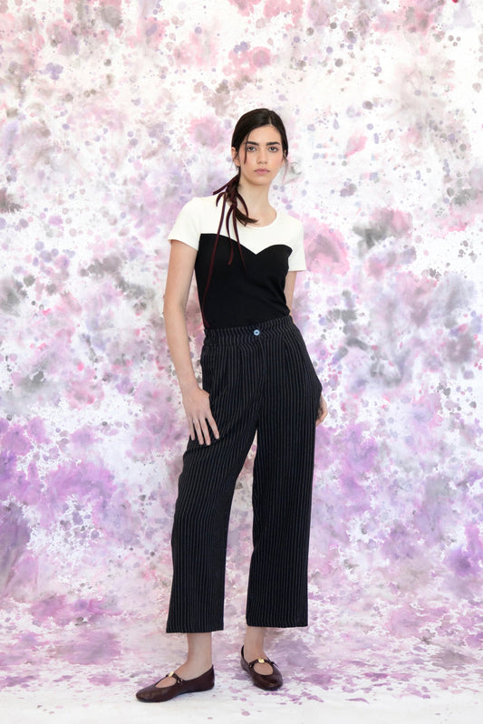 A woman wearing the Tanaisie Woven Pants from Eve Lavoie in Black Stripe, slightly cropped linen/cotton pants with wide legs, an elastic waist, classic fly closure, and slanted side pockets. She is wearing them with a black and white top and standing in front of a purple and white speckle background. 