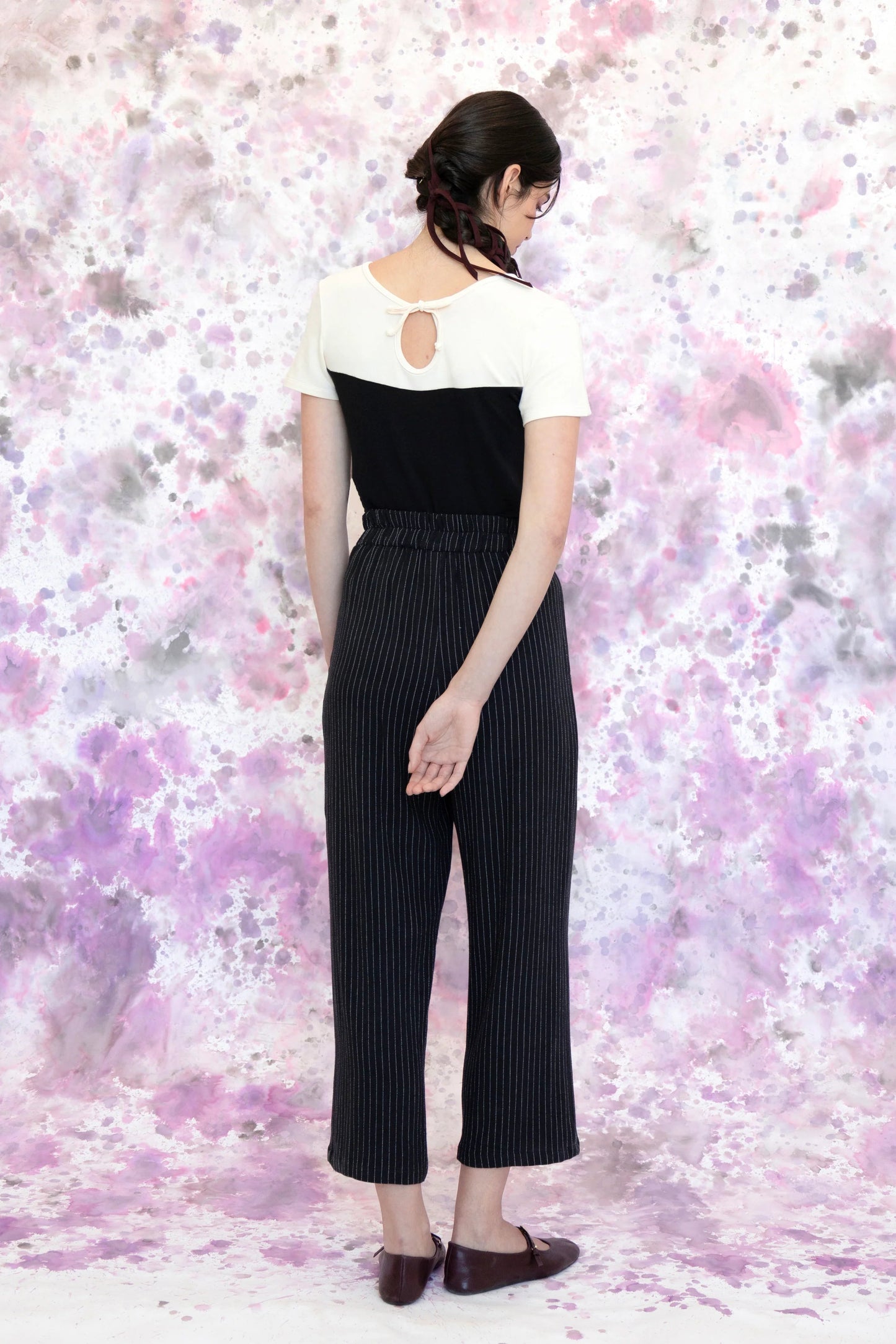 Back view of a woman wearing the Tanaisie Woven Pants from Eve Lavoie in Black Stripe, slightly cropped linen/cotton pants with wide legs, an elastic waist, classic fly closure, and slanted side pockets. She is wearing them with a black and white top and standing in front of a purple and white speckle background. 