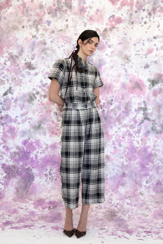 A woman wearing the Eglantine Plaid Blouse by Eve Lavoie in Black and Cream Plaid, with a mandarin collar, short puffed sleeves, rounded hem, and button front. She is wearing it with matching pants and standing in front of a purple and white speckled background. 