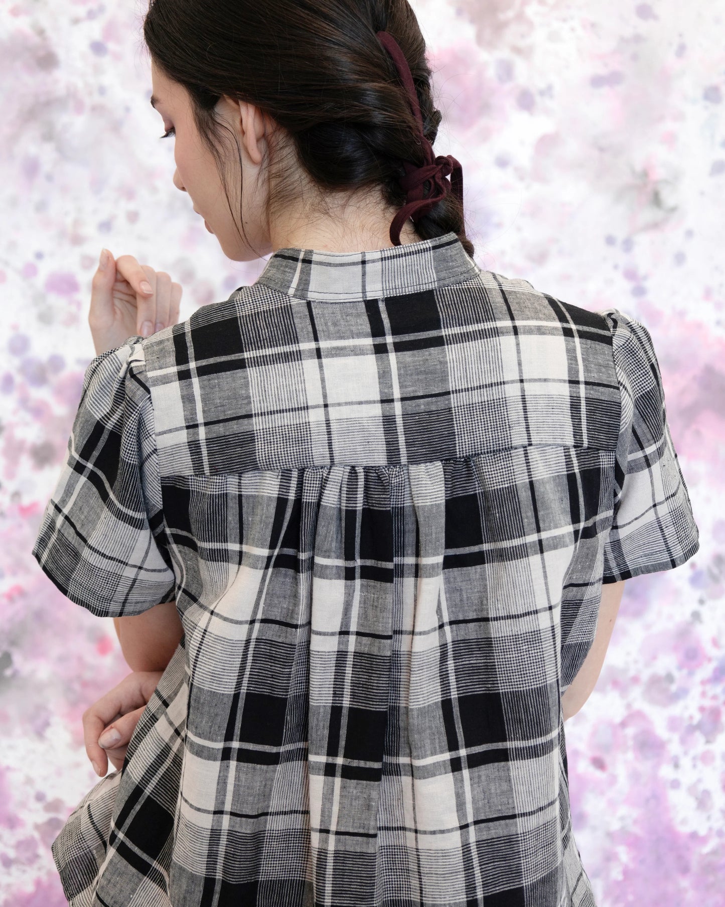 Back waist up view of a woman wearing the Eglantine Plaid Blouse by Eve Lavoie in Black and Cream Plaid, with a mandarin collar, short puffed sleeves, rounded hem, and button front. She is wearing it with matching pants and standing in front of a purple and white speckled background. 