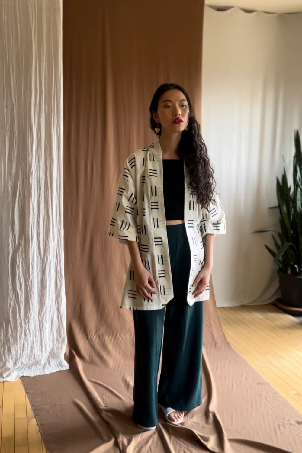 A woman wearing the Mirano Jacket by Kazak in Cream Lines print, an open Kimono-inspired jacket that falls to mid-thigh and has wide 3/4 sleeves. She is wearing it with a black top and green pants and is standing in front of a beige curtain. 