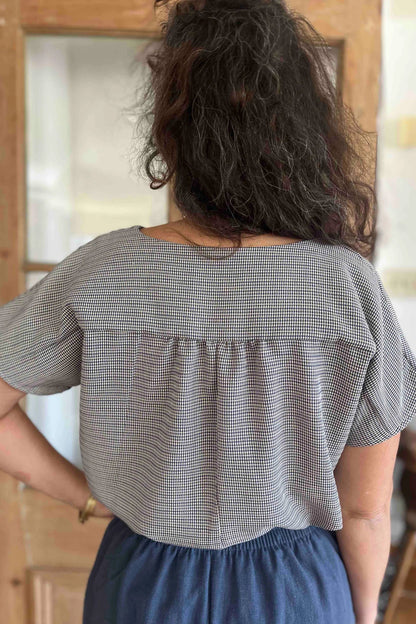 Back view of a woman wearing  the Lucca Top in Blue Check print by Kazak, a short sleeved top with a notched V-neckline and a pleated yoke at the back. She is wearing it with the Sienna Pants in Lapis, and is standing in front of a wooden door. 
