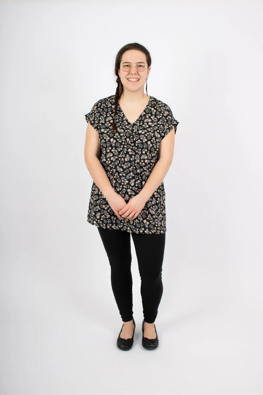 Woman wearing the Marissa Tunic by Infime in Black floral, with a v-neck placket, short extended sleeves with a cuff detail, and side slits, with black pants in front of a white background. 