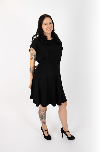 A woman wearing the Miyuki Dress by Infime in Black, with glittery fabric, a boat neck, short extended sleeves, a contrast waistband, and a flared knee-length skirt, standing in front of a white background
