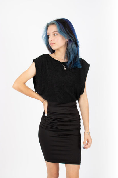 A woman wearing the Saki Top by Infime in glitter Black with a black skirt, standing in front of a white background 