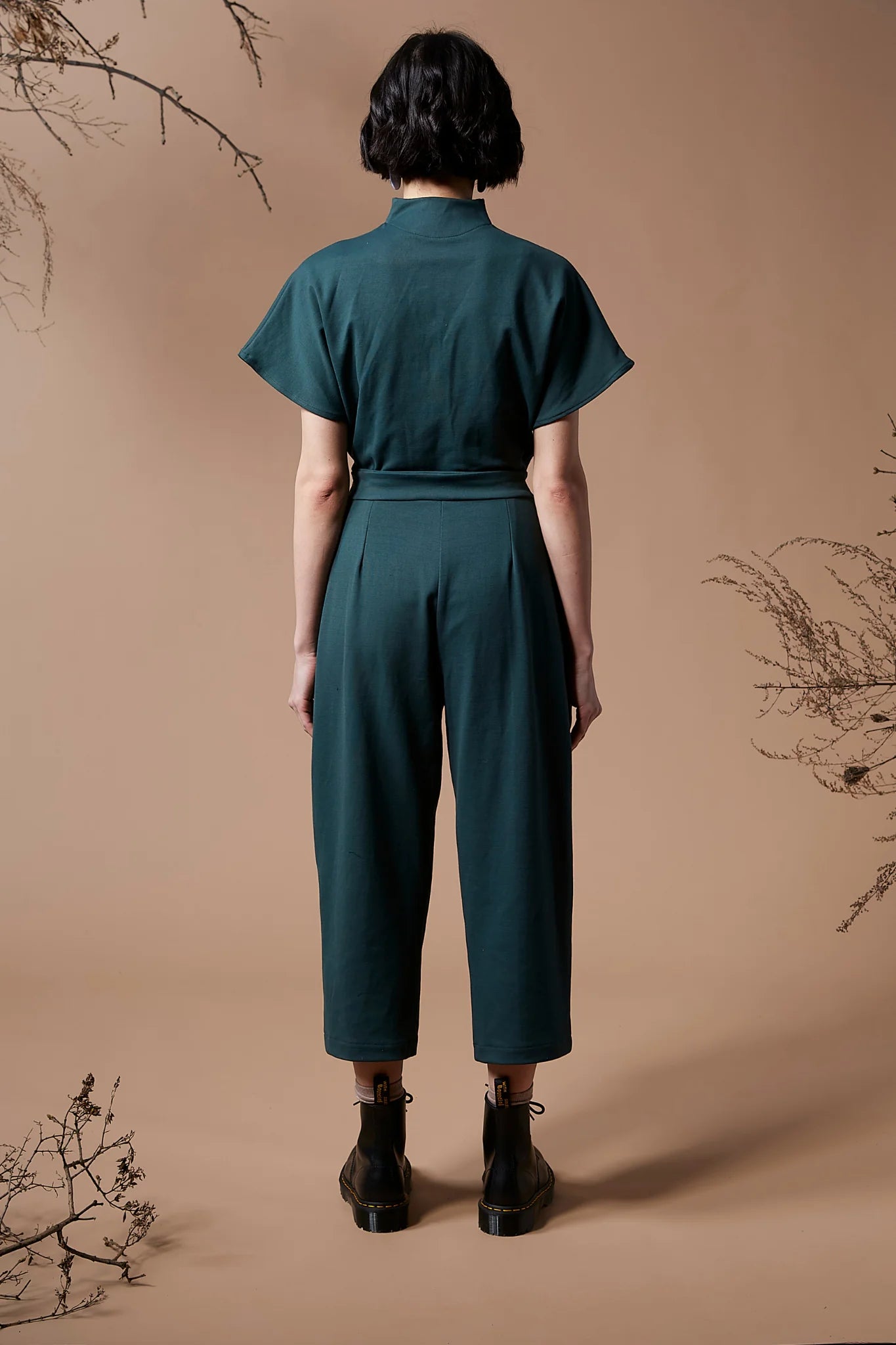 Back view of a woman wearing the Jayden Jumpsuit by Melow in Forest, standing in front of a beige background