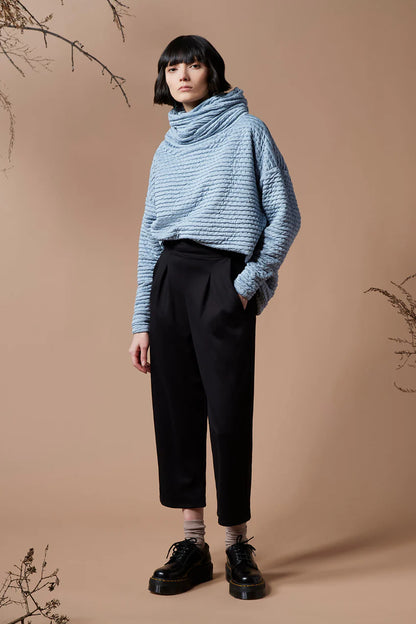 A woman wearing the Wilfred Pants by Melow in Black, with cropped legs, a pull on waist band and front pleats, with a blue sweater, standing in front of a beige background. 