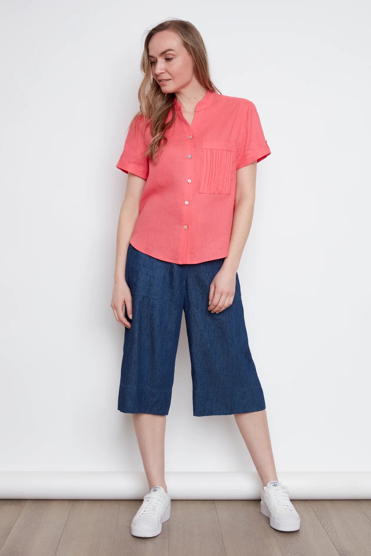 Close up view of a woman wearing the Jasmine Short Sleeve Linen Shirt by Julei in Coral, with a mandarin collar, pleated pocket, and cuff and tab sleeves. She is wearing it with denim capris and standing in front of a white background. 