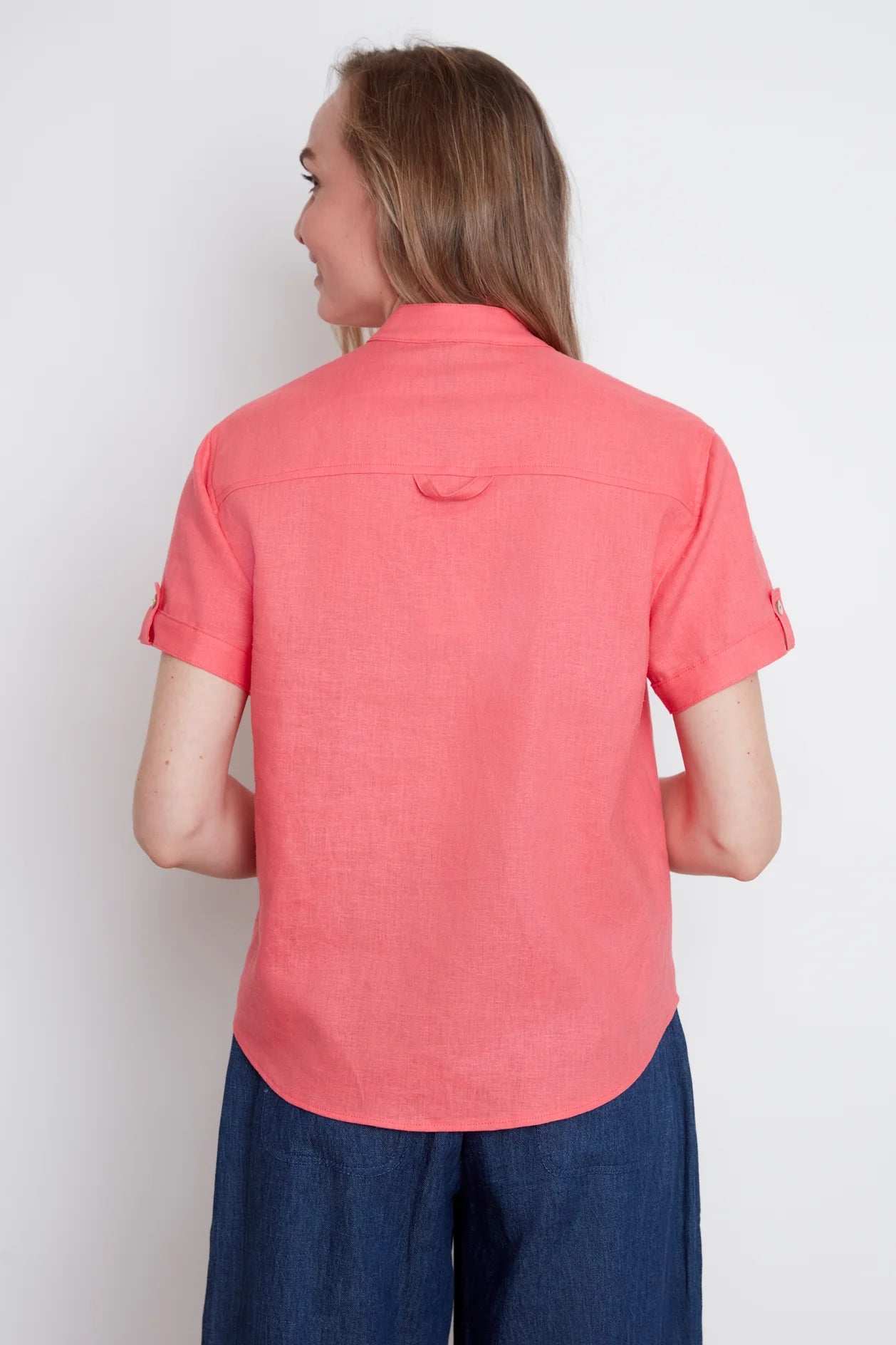 Back view of a woman wearing the Jasmine Short Sleeve Linen Shirt by Julei in Coral, with a mandarin collar, pleated pocket, and cuff and tab sleeves. She is wearing it with denim capris and standing in front of a white background. 