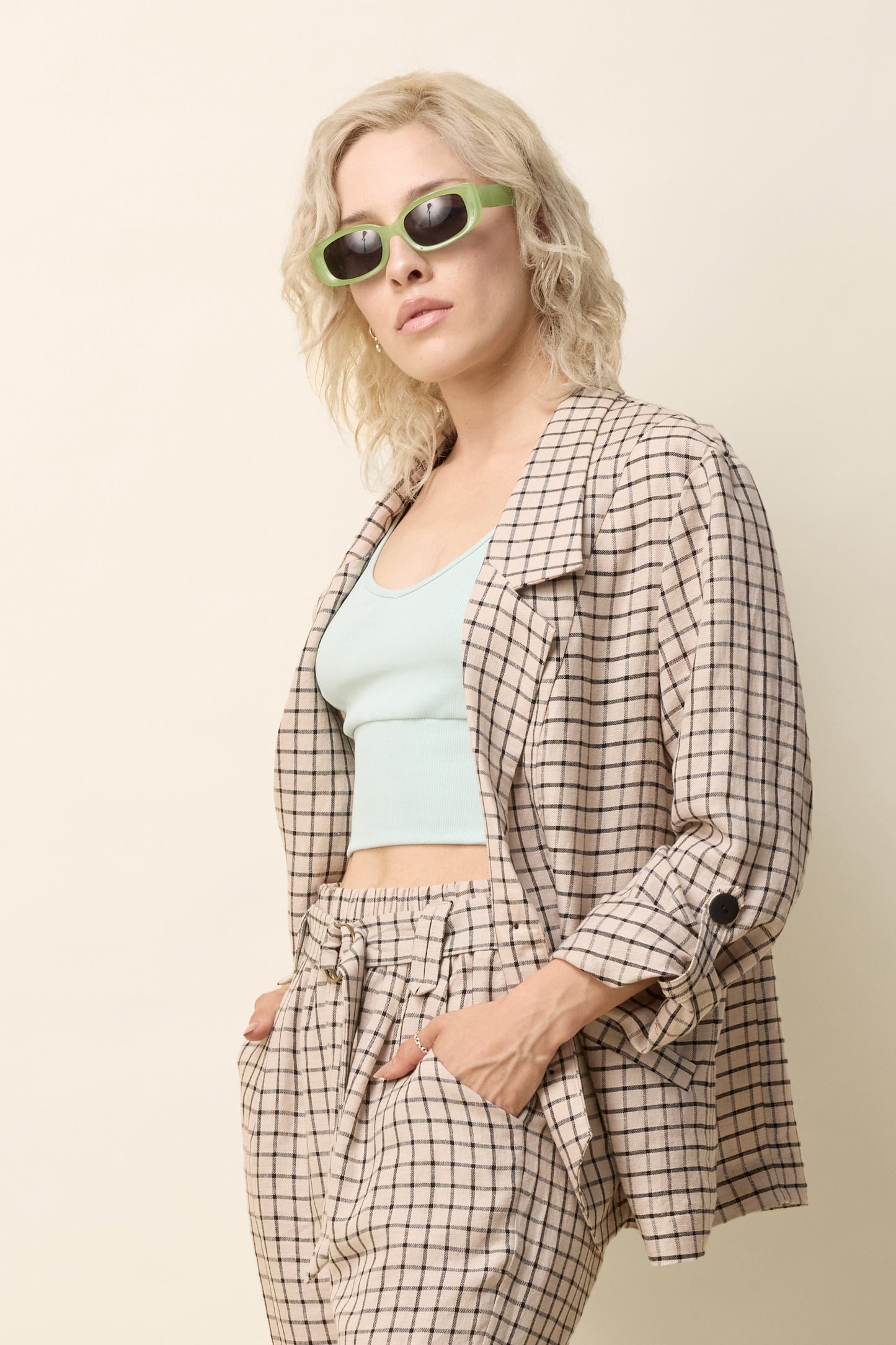 A woman wearing the Jennifer Jacket by Cokluch in Natural Utopia - a beige checkered pattern - with a wide lapel collar, two front buttons, angled front helm, and adjustable sleeves. She is wearing it with matching pants and standing in front of a beige background. 