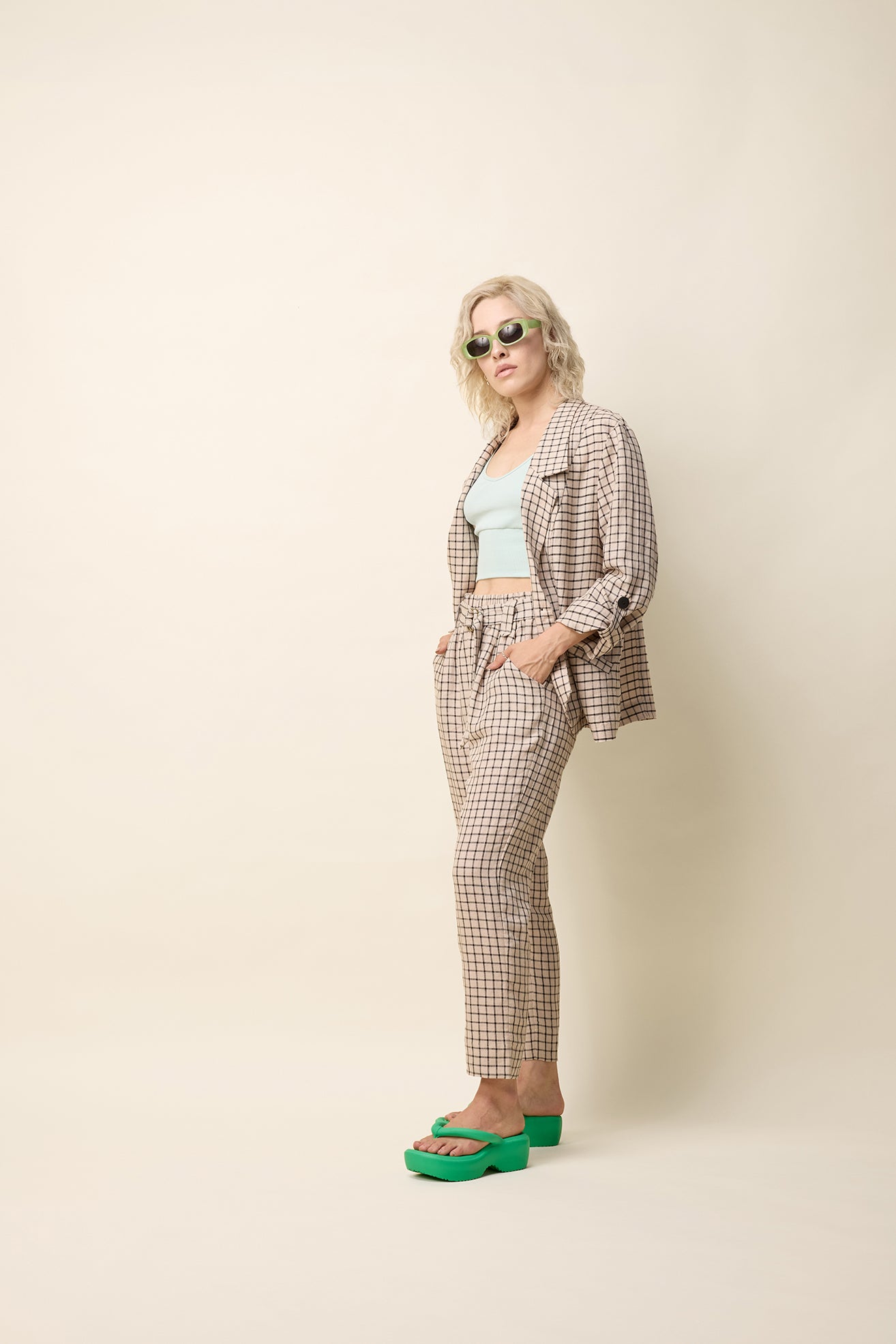 A woman wearing the Jennifer Jacket by Cokluch in Natural Utopia - a beige checkered pattern -  with a wide lapel collar, two front buttons, angled front helm, and adjustable sleeves. She is wearing it with matching pants and standing in front of a beige background. 