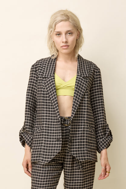 A woman wearing the Jennifer Jacket by Cokluch in Black Utopia - black checkered pattern - with a wide lapel collar, two front buttons, angled front helm, and adjustable sleeves. She is wearing it with the matching Yasmina Pants and standing in front of a beige background. 