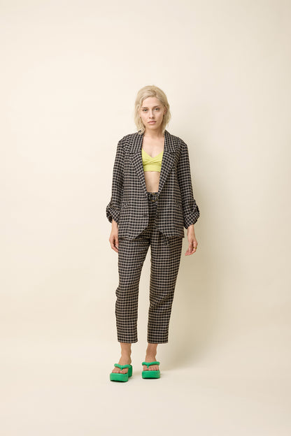 A woman wearing the Jennifer Jacket by Cokluch in Black Utopia - black checkered pattern - with a wide lapel collar, two front buttons, angled front helm, and adjustable sleeves. She is wearing it with the matching Yasmina Pants and standing in front of a beige background. 