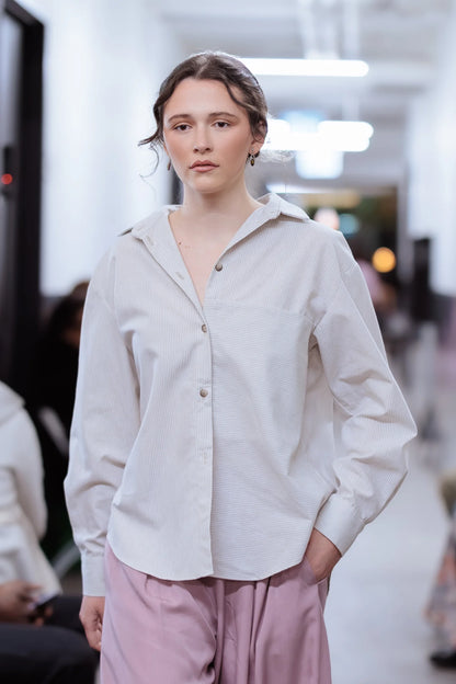 Woman wearing the Kyoto Blouse by MAS in Stripe, a classic collared button down with a cross-pleat at the back. She is wearing it open with rousers and walking through crowd.
