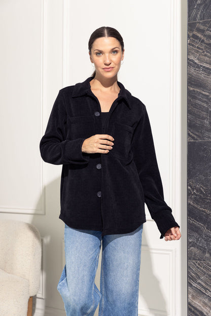 A woman wearing the Joplin Jacket by Cherry Bobin in Black, a shirt jacket in corduroy fabric with a collar and button front and two breast pockets. She is wearing it over jeans and a black shirt and is standing in front of a grey wall. 