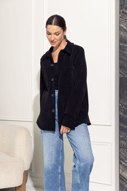 A woman wearing the Joplin Jacket by Cherry Bobin in Black, a shirt jacket in corduroy fabric with a collar and button front and two breast pockets. She is wearing it over jeans and a black shirt and is standing in front of a grey wall. 