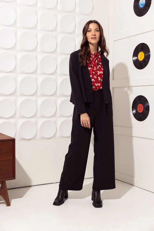 A woman wearing the Santana Pants by Cherry Bobin in Black, classic straight cut trousers with a fly closure, side pockets, and front box pleats. She is wearing them with a red top and black blazer and is standing in front of a white wall with vinyl records on it.