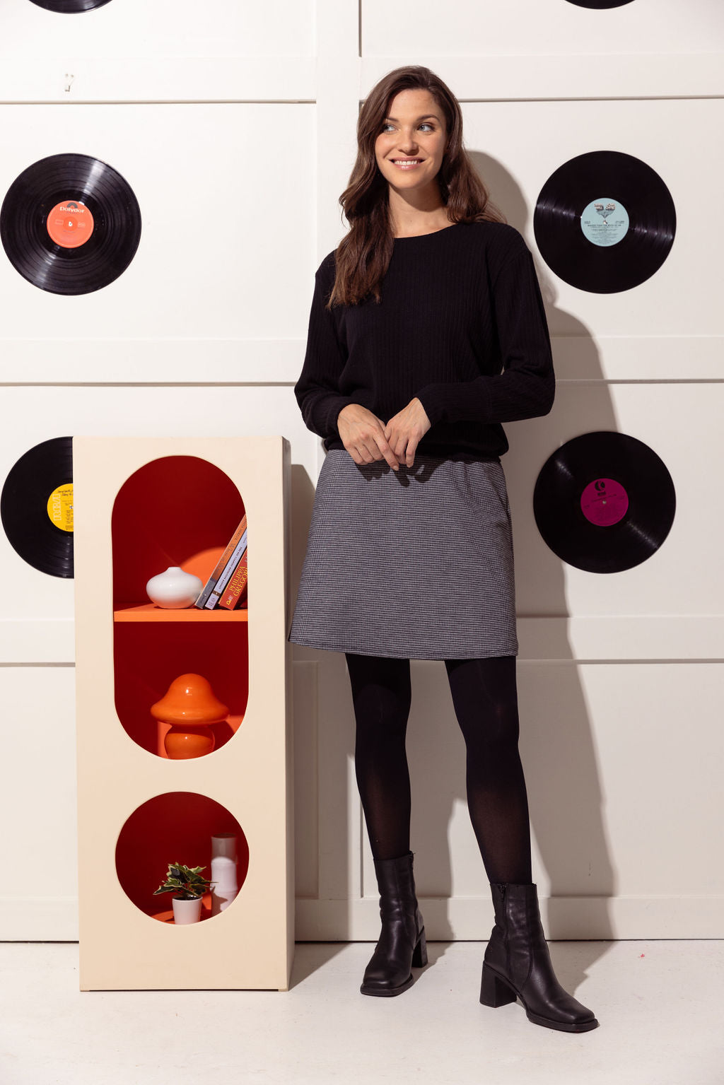 A woman wearing the Dylan Skirt by Cherry Bobin in Houndstooth, a short A-line skirt with an elastic waistband, with a black turtleneck. She is standing in front of a white wall with vinyl records hanging on it. 