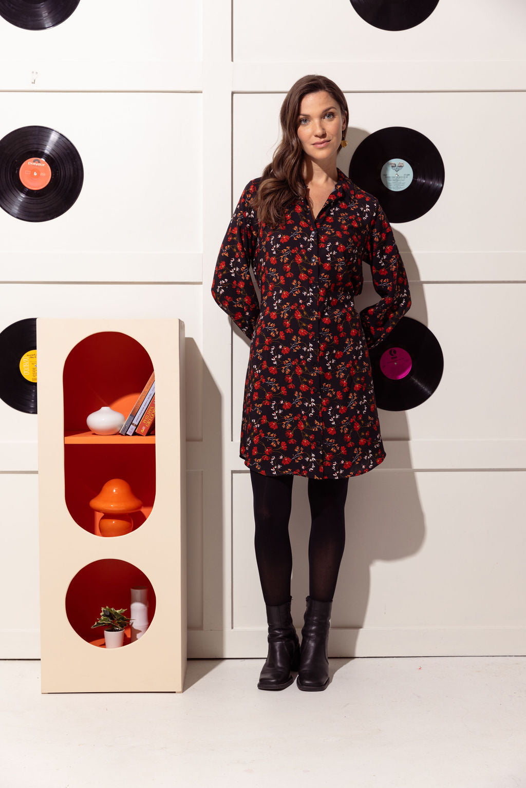 A woman wearing the Alanis Dress by Cherry Bobin in Black with Red Flowers, a classic shirt dress with a collar, button front, and tie belt and a rounded above the knee hemline. She is wearing it with black tights and is standing in front of a white wall with black vinyl records hanging on it. 