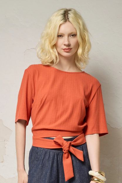 Waist up view of a woman wearing the Kali Top by Cokluch in Fiesta, a ribbed knit top with elbow-length sleeves, an integrated built, and an opening at the lower back. She is wearing it with blue pants and standing against a neutral background.