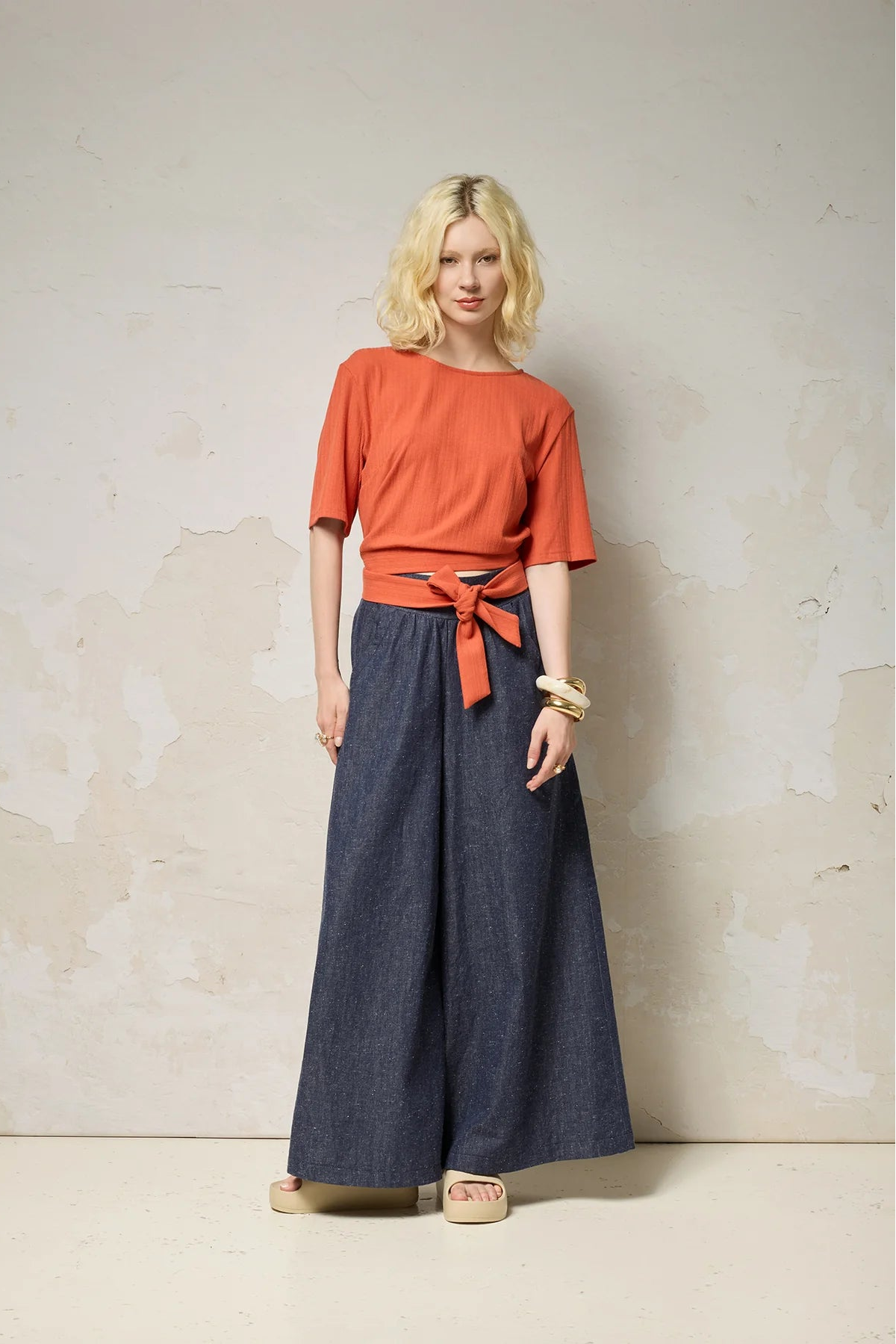 A woman wearing the Kali Top by Cokluch in Fiesta, a ribbed knit top with elbow-length sleeves, an integrated built, and an opening at the lower back. She is wearing it with blue pants and standing against a neutral background