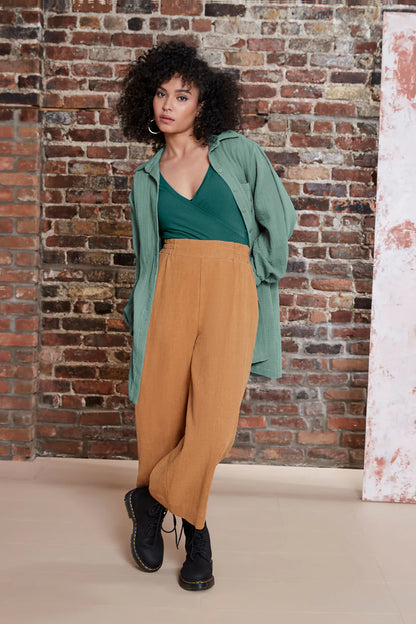 A woman wearing the Kurt Pants by Melow in Honey, featuring wide cropped legs, a flat-front elastic waist band, and slant pockets.  She is wearing them with a green top and standing in front of a brick wall. 