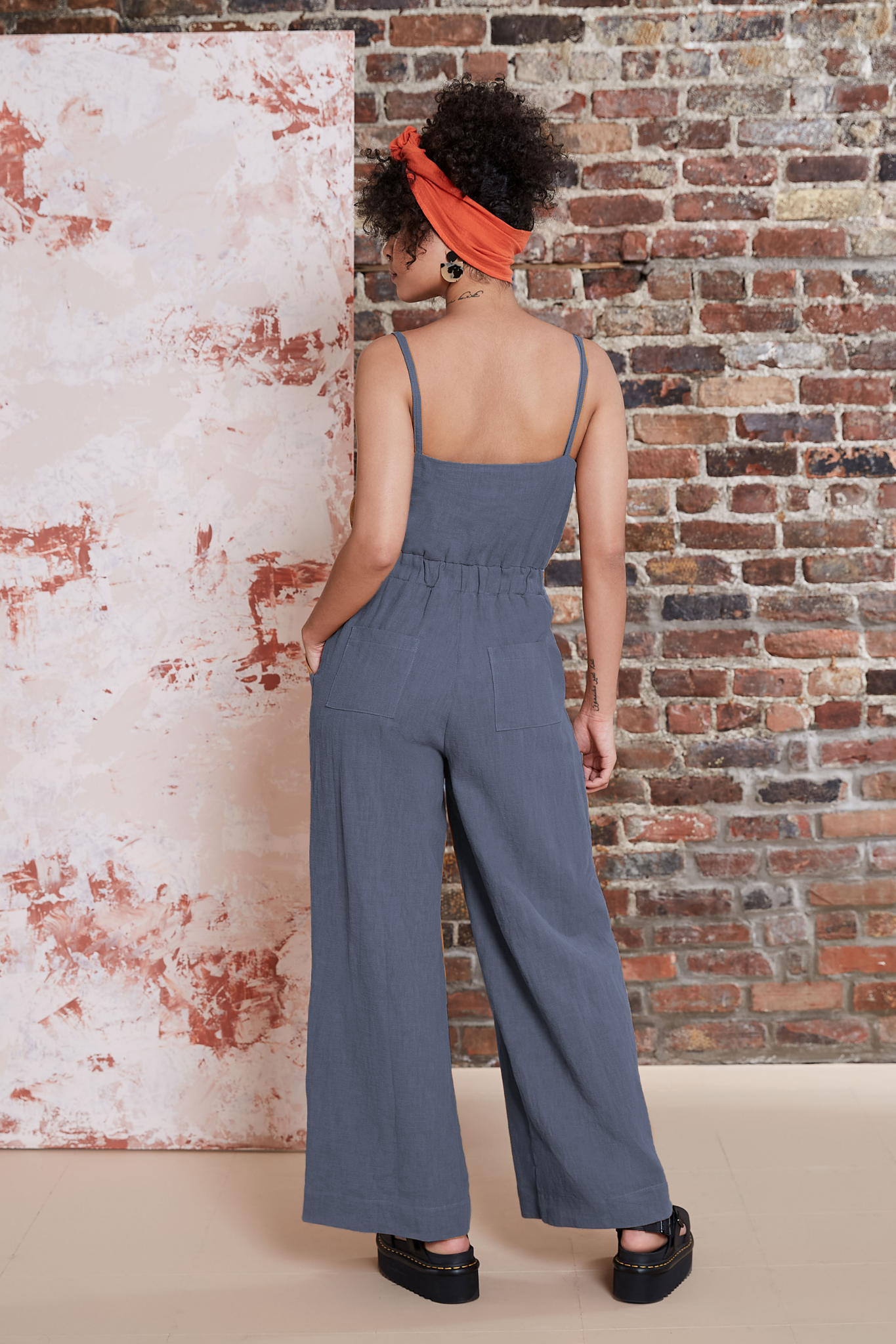 Back view of a woman wearing the Kasim Jumpsuit by Melow in Slate, featuring spaghetti straps, an elastic waist, wide full-length legs, and side pockets. She is standing against a brick wall. 