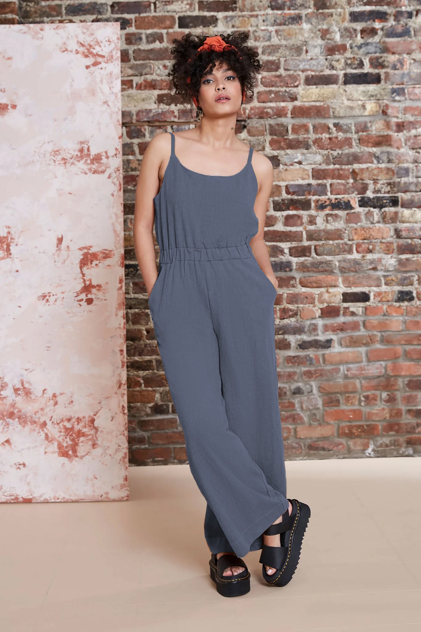 A woman wearing the Kasim Jumpsuit by Melow in Slate, featuring spaghetti straps, an elastic waist, wide full-length legs, and side pockets. She is standing against a brick wall. 