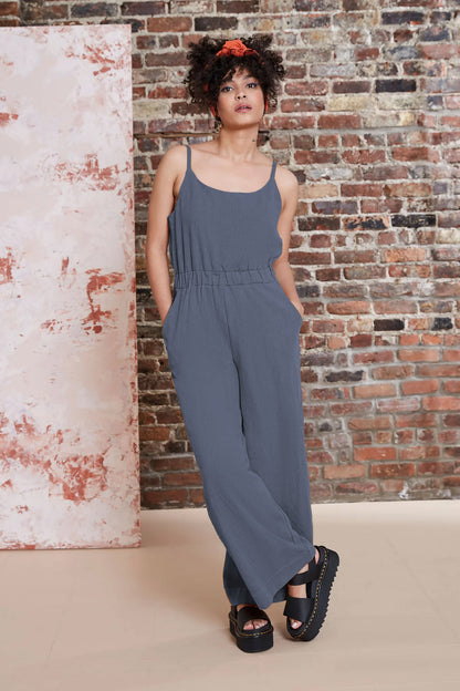 A woman wearing the Kasim Jumpsuit by Melow in Slate, featuring spaghetti straps, an elastic waist, wide full-length legs, and side pockets. She is standing against a brick wall. 