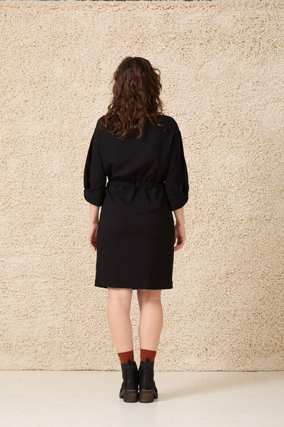 Back view of a woman wearing the Kate Dress by Cokluch in Black, featuring a drawstring waist, 3/4 sleeves with a twist detail at the cuffs, shoulder darts, and a mid-thigh length. She is standing in front of a speckled beige background. 