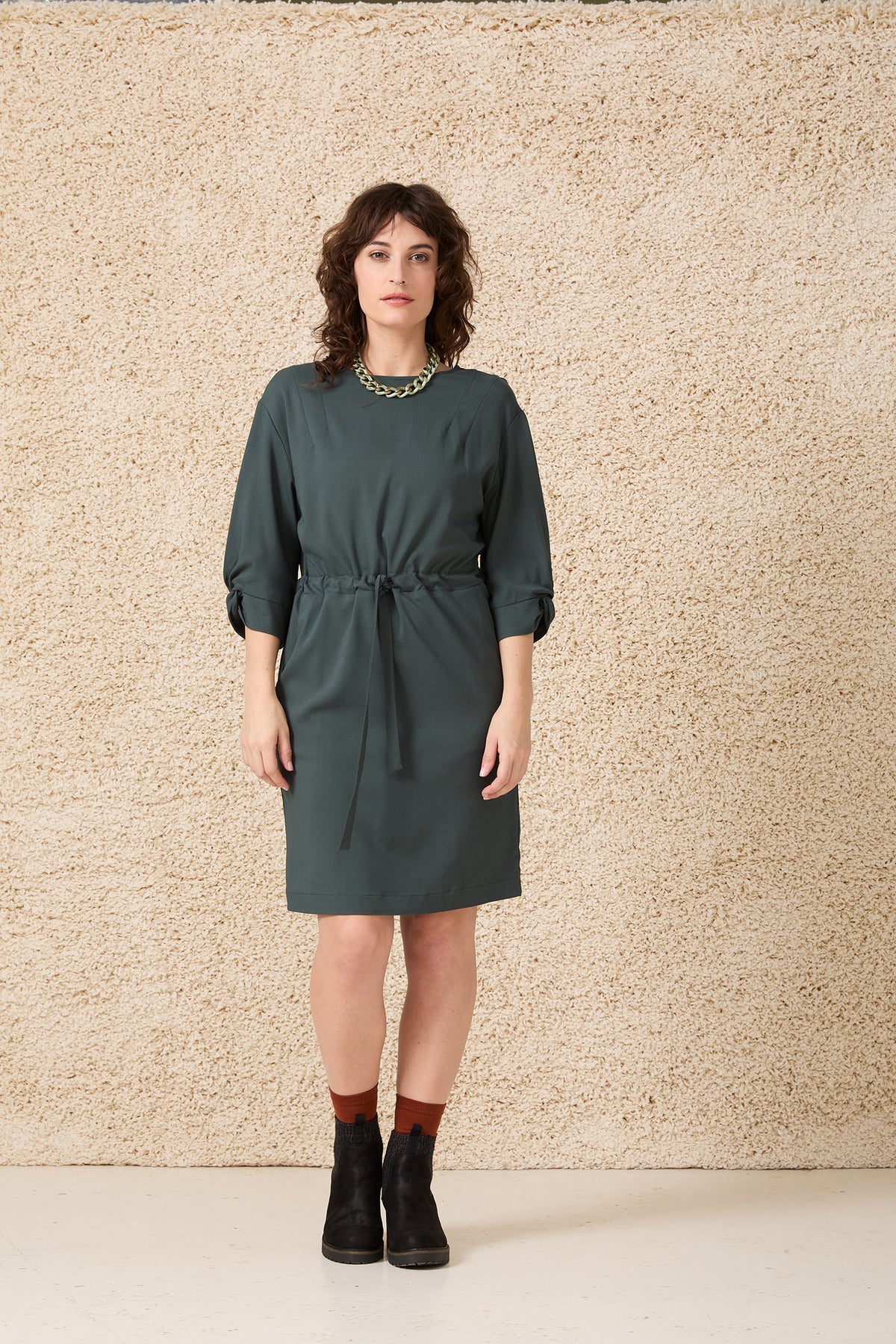 A woman wearing the Kate Dress by Cokluch in Cilantro, featuring a drawstring waist, 3/4 sleeves with a twist detail at the cuffs, shoulder darts, and a mid-thigh length. She is standing in front of a speckled beige background. 