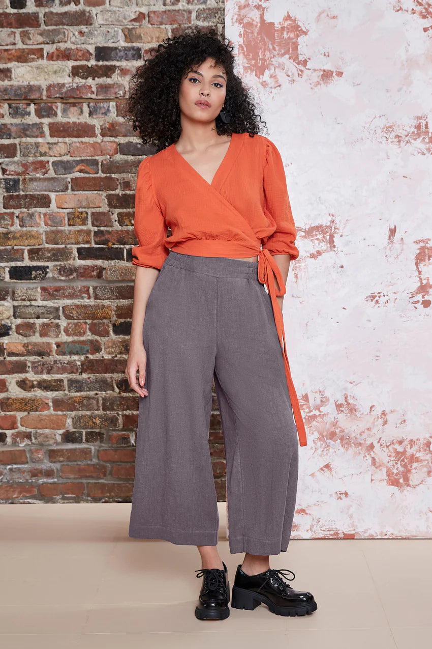 A woman wearing the Kurt Pants by Melow in Slate, featuring wide cropped legs, a flat-front elastic waist band, and slant pockets.  She is wearing them with an orange top and standing in front of a brick wall. 